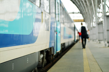 blue and grey train with metal construction, platform and person in the background, partly blurred