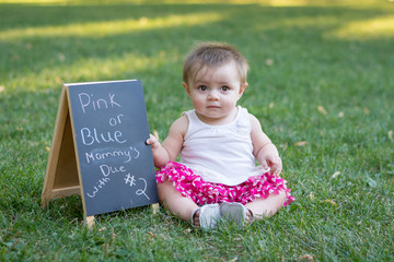 7 month old girl sits in the grass with a pregnancy anouncement sign
