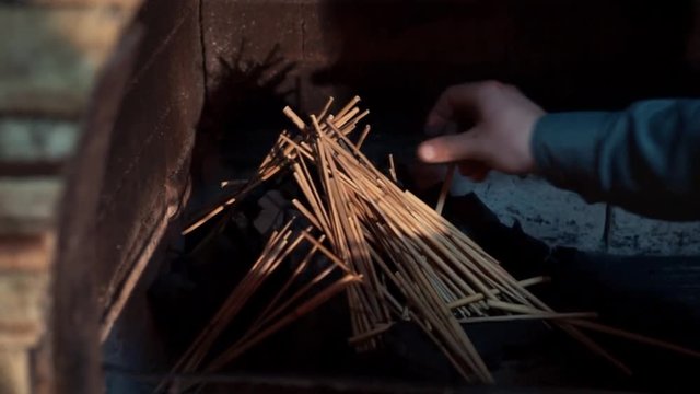 Male Hand In Grey Shirt Black Vest Puts Wooden Kindling Sticks Over Charcoal Pile In Indoor Fireplace, Making Fire