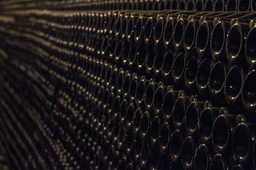 Many bottles of champagne in a dark wine cellar under the ground lie in even rows under the light of a yellow lamp.