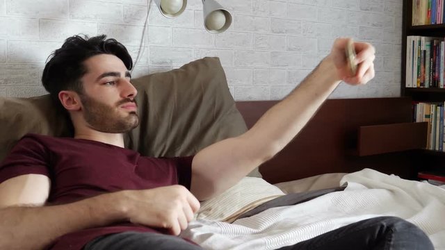 Young attractive man taking selfie photo on the phone while lying on the bed at home