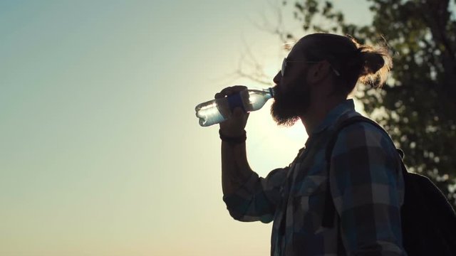 Side view of stylish man with backpack drinking water from bottle on background of sunlight and nature. Slow motion.