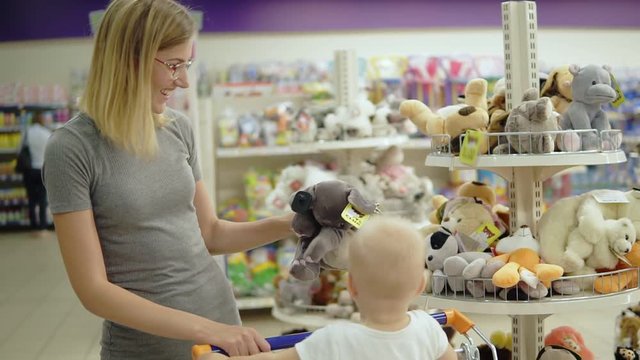 Smiling mother showing her child a toy elephant in a toy section for children in the supermarket while her cute little child sitting in a shopping cart. Family shopping
