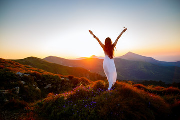 Hiker woman standing with hands up achieving the top. Girl welcomes a sun. Conceptual design. Successful woman hiker open arms on sunrise mountain top. Girl in long white dress in the mountains