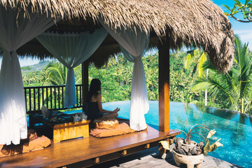 Girl meditating by the pool on a background of palms, Bali, Indonesia.The girl sits in a gazebo at a bali in a lotus pose and meditates with a view of the palms and the pool. Breakfast in Bali