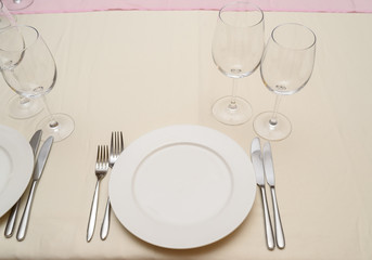 Dinner place setting in restaurant. Empty white plate with silver fork and knife, transparent wineglasses on beige tablecloth, free space for text. Elegant table setting for dining in restaurant