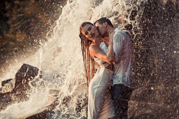 Young enamored couple hugs and kisses under spray of waterfall.