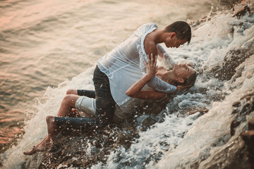 Young enamored couple lies on rock and hugs under spray of waterfall.