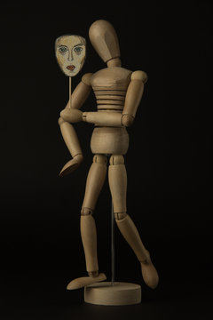 Wooden doll on hinges holds a mask in hands and covers her face on a black background