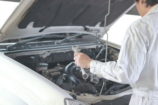Side view of automotive mechanic in white uniform with wrench diagnosing engine under hood of car at the repair garage.