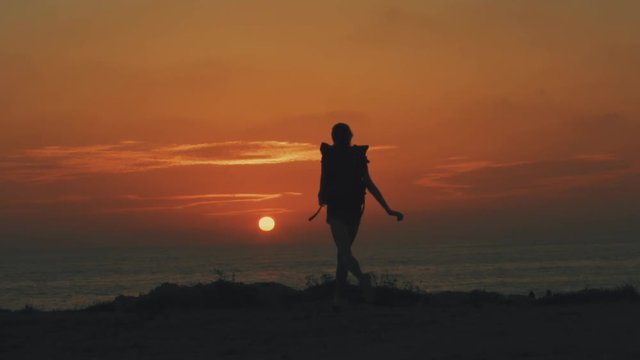 A young traveler girl with a backpack runs to the sea and the orange sun at sunset and dawn, puts her hands up, sunlight flashes, Silhouette of a girl, 100 frames per second, slow motion video.