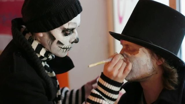 Young Woman Applying Makeup Onto Man's Face At Halloween