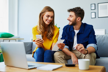 Portrait of a cute young couple sitting on sofa at home and managing budget using phone and laptop