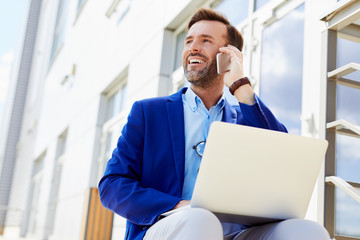 Portrait of laughing middle-aged businessman talking on phone and working on laptop while sitting outside - Powered by Adobe