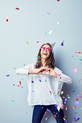 Beautiful laughing young woman celebrating birthday making heart gesture with birthday hat sunglasses and confetti