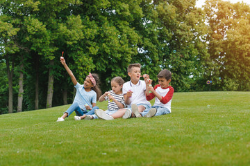 Happy multiethnic children sitting on green meadow and blowing soap bubbles in park