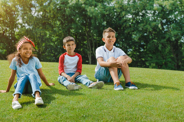 Fototapeta premium Cute smiling multiethnic kids sitting together on green lawn in park