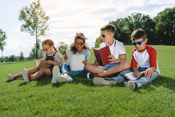 Cute multiethnic children in sunglasses reading books on green meadow