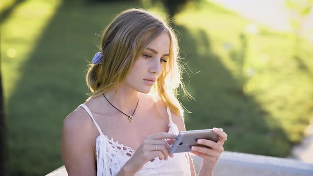 pensive blonde with light eyes, looking at cellphone, touching screen in park, sunny summer weather