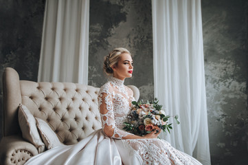 Beautiful bride in cream wedding lace dress with wedding bouquet with orange and white flowers. Studio, gray background, modern, sofa, cosmetics, makeup. 