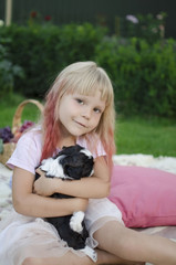 Little blond girl sitting on the cover and hugging black and white shih tzu puppy. Cute girl behind the pink pillows. 