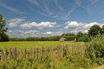 Fototapeta na wymiar Field with wooden fence. Blue sky with delicate white clouds. Polish village view.