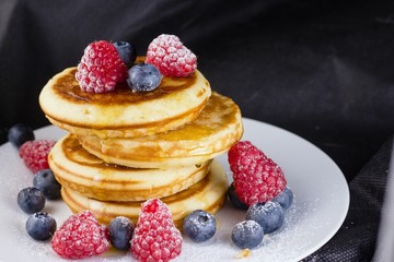 Stack of pancakes with raspberries and blueberries  coated with powdered sugar on white plate and black background
