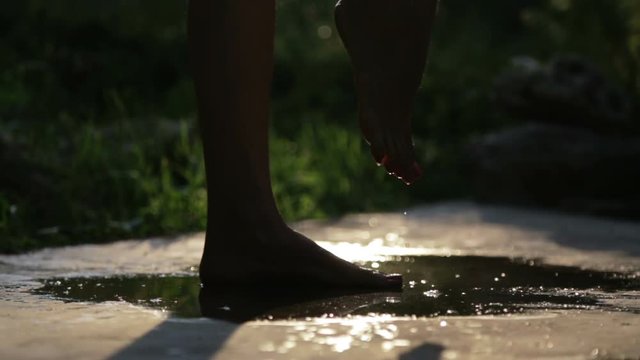 Female feet barefoot in a puddle in backlight. Silhouette of female legs in a puddle with water at sunset.
