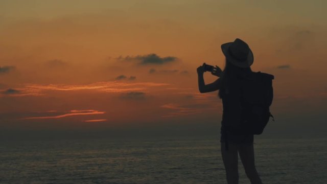 A young girl traveler with a backpack and hat on her head takes a picture on mobilephone of sea on the sunset in Cyprus, makes photos of the orange sun and sky, Silhouette of a girl, 100 frames per