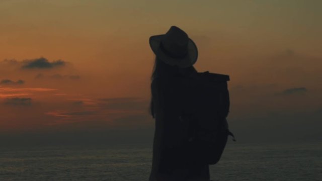 A young girl traveler with a backpack and hat on her head goes to the sea and takes pictures on mobilephone of the sunset dawn in Cyprus, makes selfies on the sea against a background of orange sun