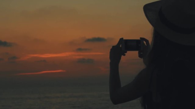 Sexy young traveler woman with a backpack and straw hat on head takes pictures on mobilephone of orange sky and sea on sunset and dawn on the sea in Cyprus, close up, Silhouette of a girl, 100 frames