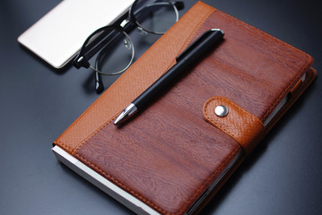 Leather Management Diary , Ball Pen , Black Eye Glasses , And Smartphone Over Black Background
