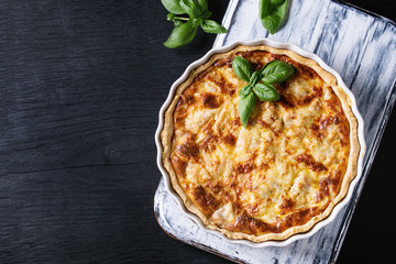 Baked homemade quiche pie in white ceramic form served with fresh greens on white serving board on...