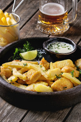 Traditional british fast food fish and chips. Served with white cheese sauce, lime, parsley in terracotta tray, glass of lager beer over old wooden plank background. Close up