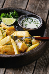 Traditional british fast food fish and chips. Served with white cheese sauce, lime, parsley, fork in terracotta tray over old wooden plank background. Close up