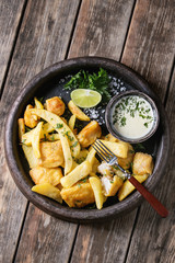 Traditional british fast food fish and chips. Served with white cheese sauce, lime, parsley, fork in terracotta tray over old wooden plank background. Top view with space