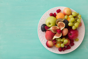 Top view on assortment of juicy fruits on white plate and turquoise wooden table background. Organic raspberries, apricots, apples, figs,plums, grapes - summer dessert or snack. healthy eating concept