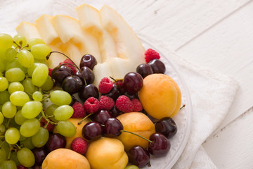 Assortment of juicy fruits on white plate and white table background. Organic raspberries, apricots, melon, cherries - summer dessert or snack. healthy eating concept.
