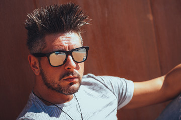 Portrait of handsome young man in casual clothing with sunglasses sitting on wood bench, looking away to a side. Color Toned Image. Concept urban man.