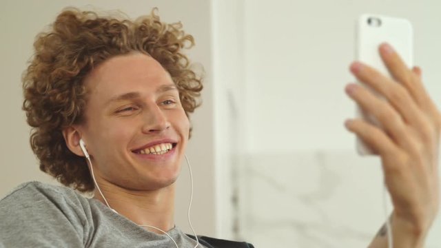 Young cheerful man in headphones making videocall on smartphone and talking to somebody at home