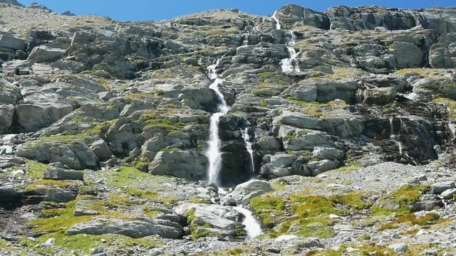 Waterfalls flowing in idyllic uncontaminated environment crossing green meadows and boulders on the Alps in summer. Slow motion