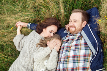 Top view of friendly happy family lying on green grass, embracing and having pleasant feelings. Little girl closing her face with hands while lying near her affectionate parents, resting on meadow