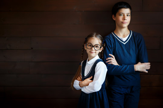 Attractive boy keeping his hands crossed while standing near his little sister in big glasses and school uniform. Positive pupils isolated over blank studio background. School, children, learning