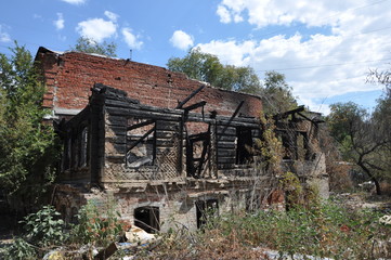 Abandoned burnt house of old architecture with ruined wooden and brick walls and charry logs, all among green trees, bushes and garbage. Big clouds in the sky.
