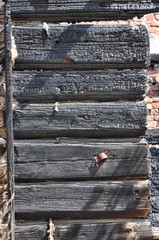 Close-up view of charry log ends of a burnt wooden house against a red brick wall.

