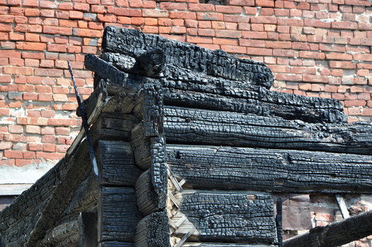 Close-up view of a charry corner of a burnt wooden house against a red brick wall.
