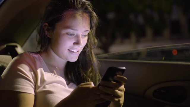 Teen Girl Plays On Her Smartphone In Back Seat Of Car During A Red Light, At Night