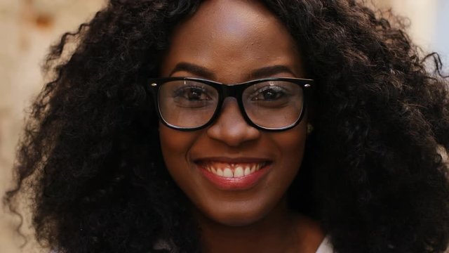 Close up portrait of beatiful curly hair african woman in the glasses smiling on the camera on the street background.