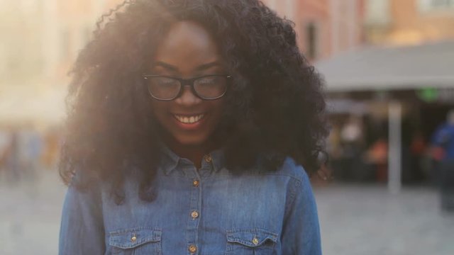 Close up shot of happy woman in the glasses using smart phone for chatting, looking to the camera and smiling on the street during sunset.
