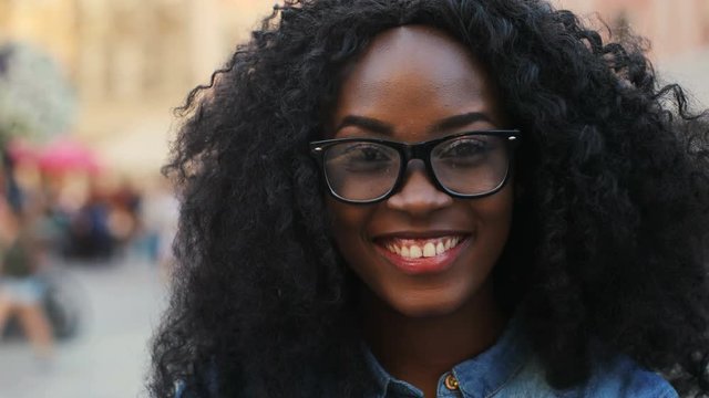 Close up portrait of attractive curly hair african woman in the glasses smiling on the camera on the street background.